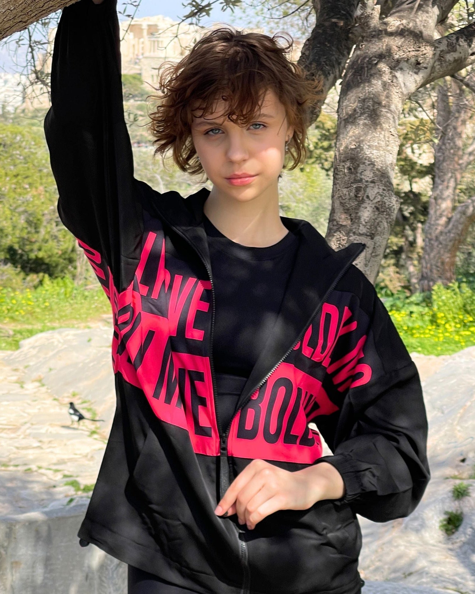 A person standing outdoors wearing a black jacket with pink text on it, raised hand holding the jacket away from the body.