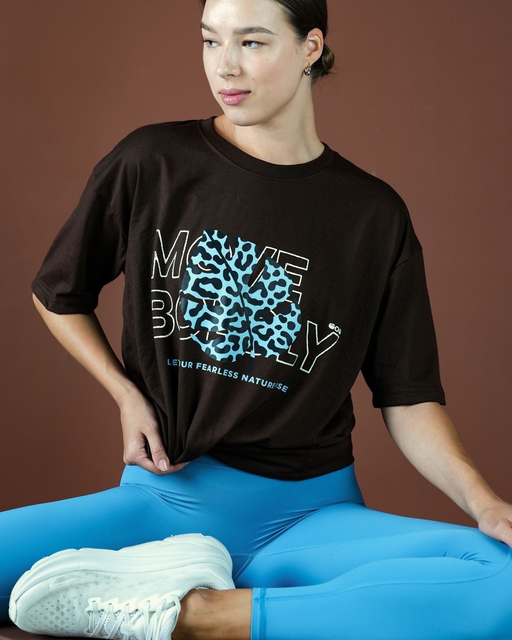 Person wearing a black t-shirt with a graphic design and blue leggings sitting on a brown background
