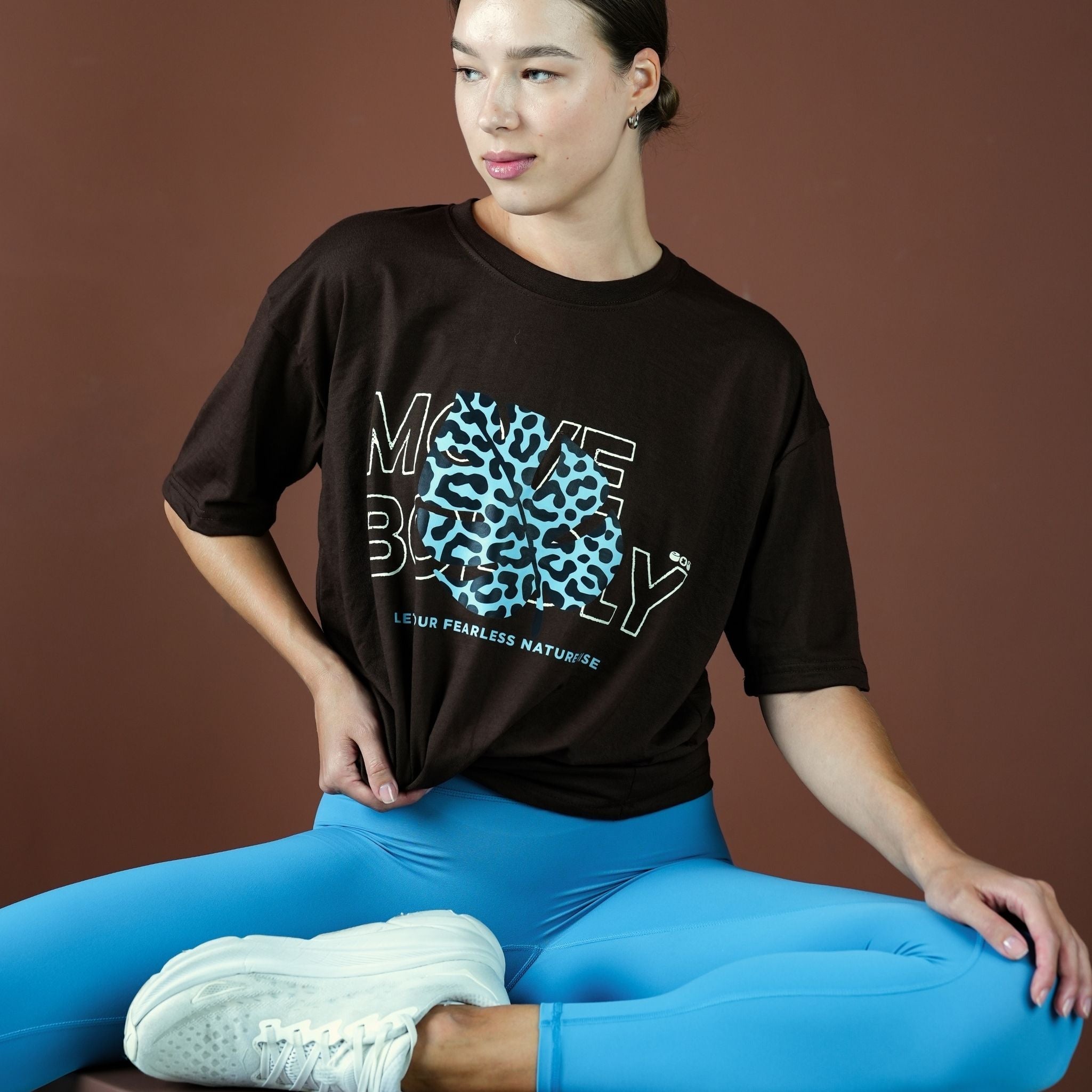 Person wearing a black t-shirt with a graphic design and blue leggings sitting on a brown background