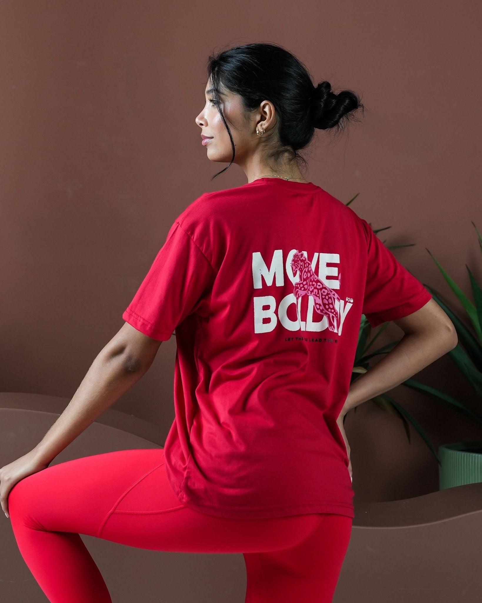 Person wearing a red 'MOVE BODY' t-shirt sitting on a couch with a plant in the background.