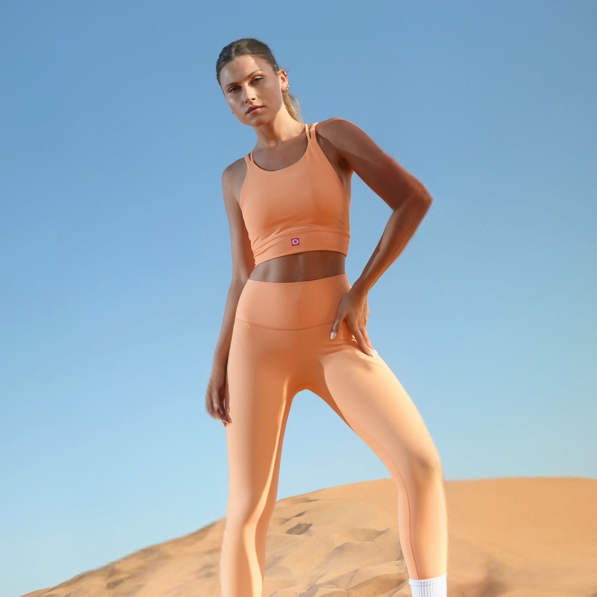 Woman in orange athletic wear standing on a sand dune with a clear blue sky.