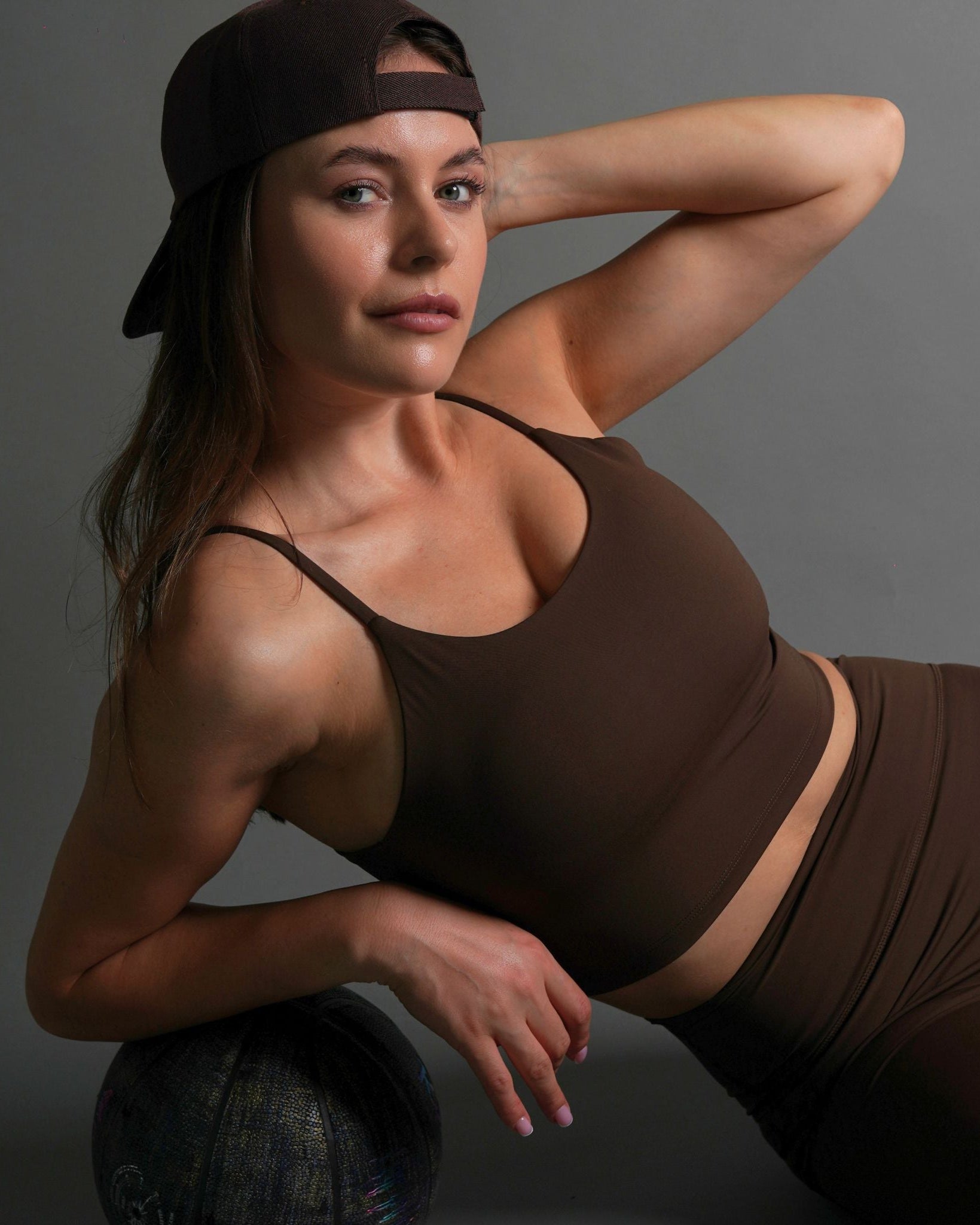 A woman wearing a brown tank bra with skinny straps, paired with matching brown leggings. She has a cap on, tilting her head to the side, and is seated against a neutral background.