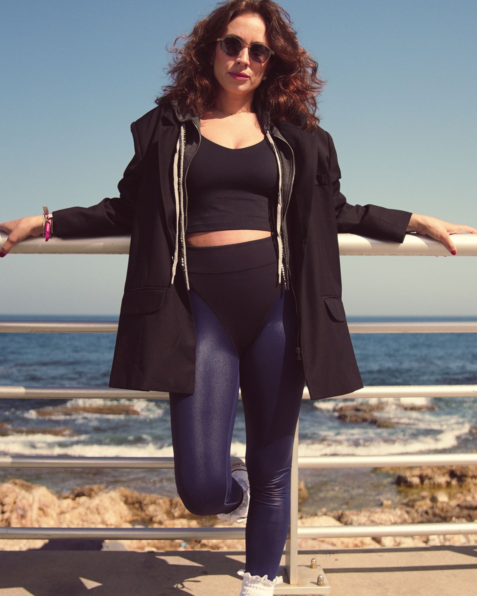 Woman in black coat and leggings standing by a railing with ocean view