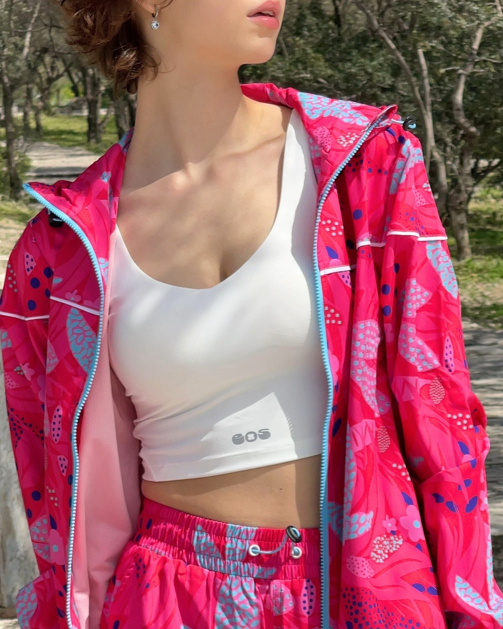 A woman wearing a white bralette with the brand name 'SOS' visible on the front, paired with a bright pink jacket and matching pants with a floral pattern.