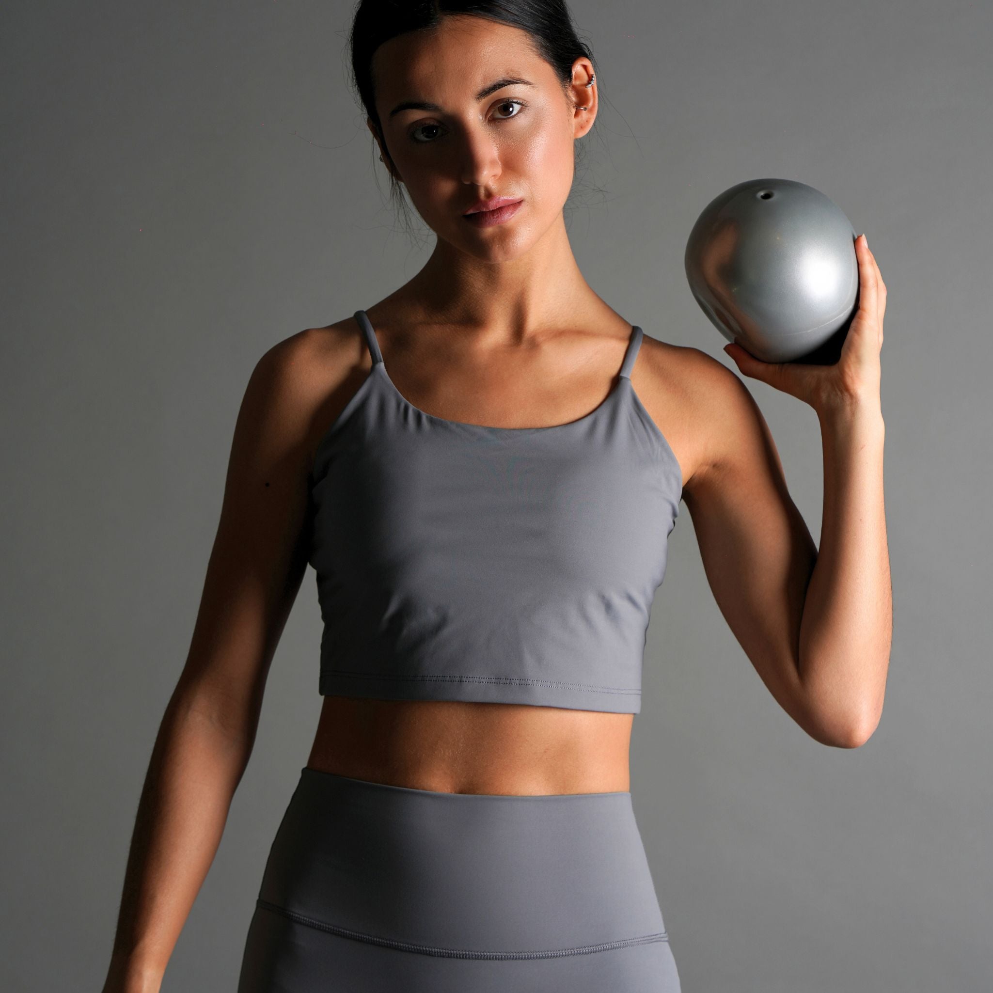 A woman wearing a gray tank bra top with skinny straps, paired with matching leggings. She is holding a exercise ball and posing against a neutral background.