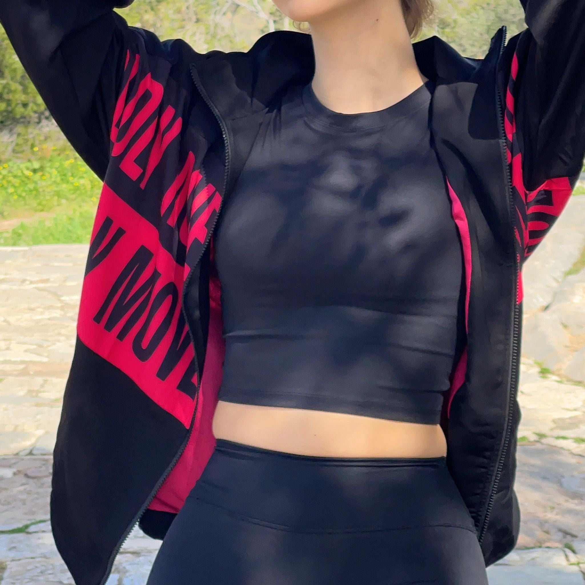 A person standing outdoors wearing a black jacket with pink text on it, raised hand holding the jacket away from the body.