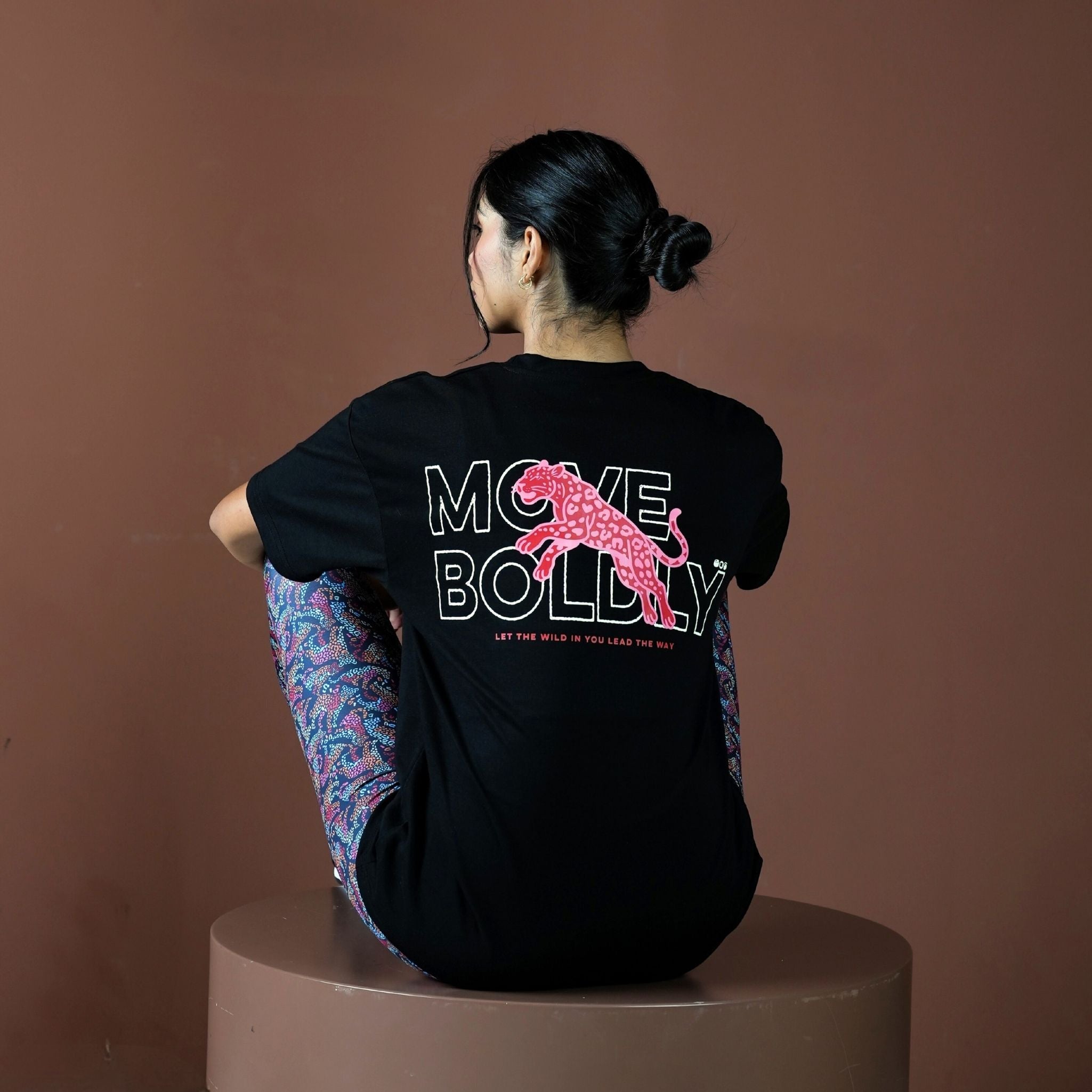 Person wearing a black t-shirt with 'Move Boldly' text and pink graphic on a brown background