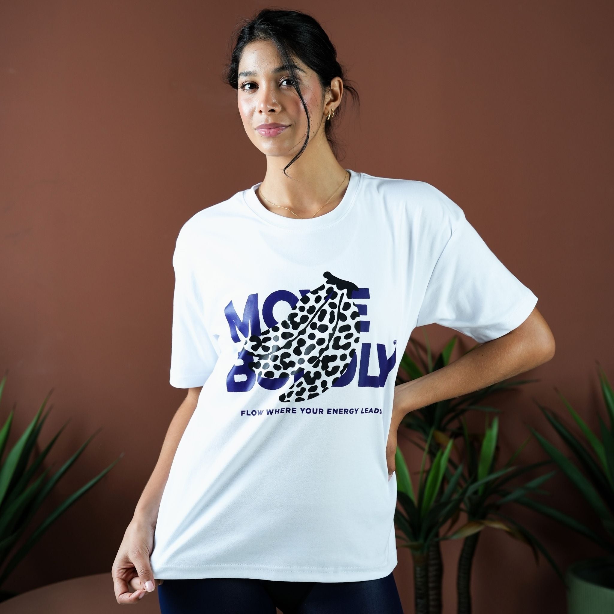 Woman wearing a white t-shirt with a graphic design and text, standing against a brown background with plants.