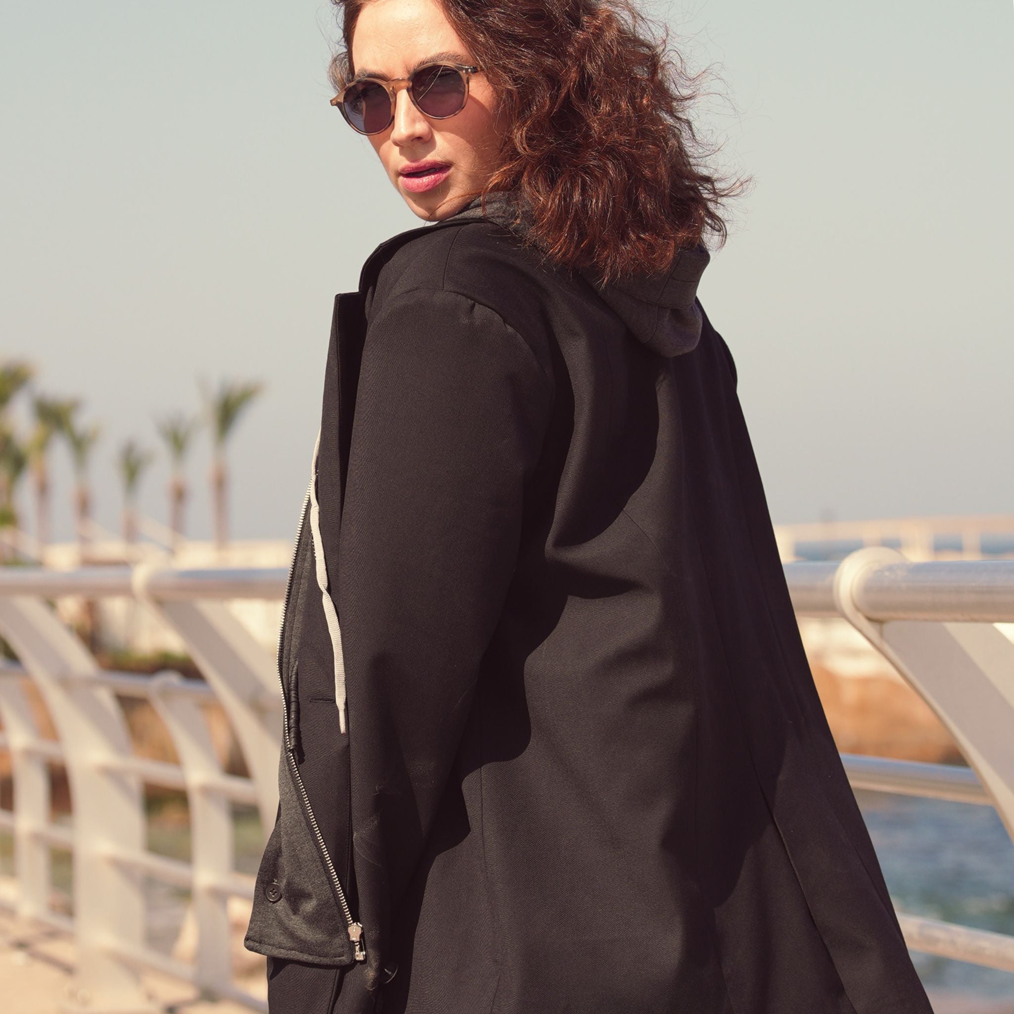 Woman wearing a black coat and sunglasses by a waterfront with palm trees in the background