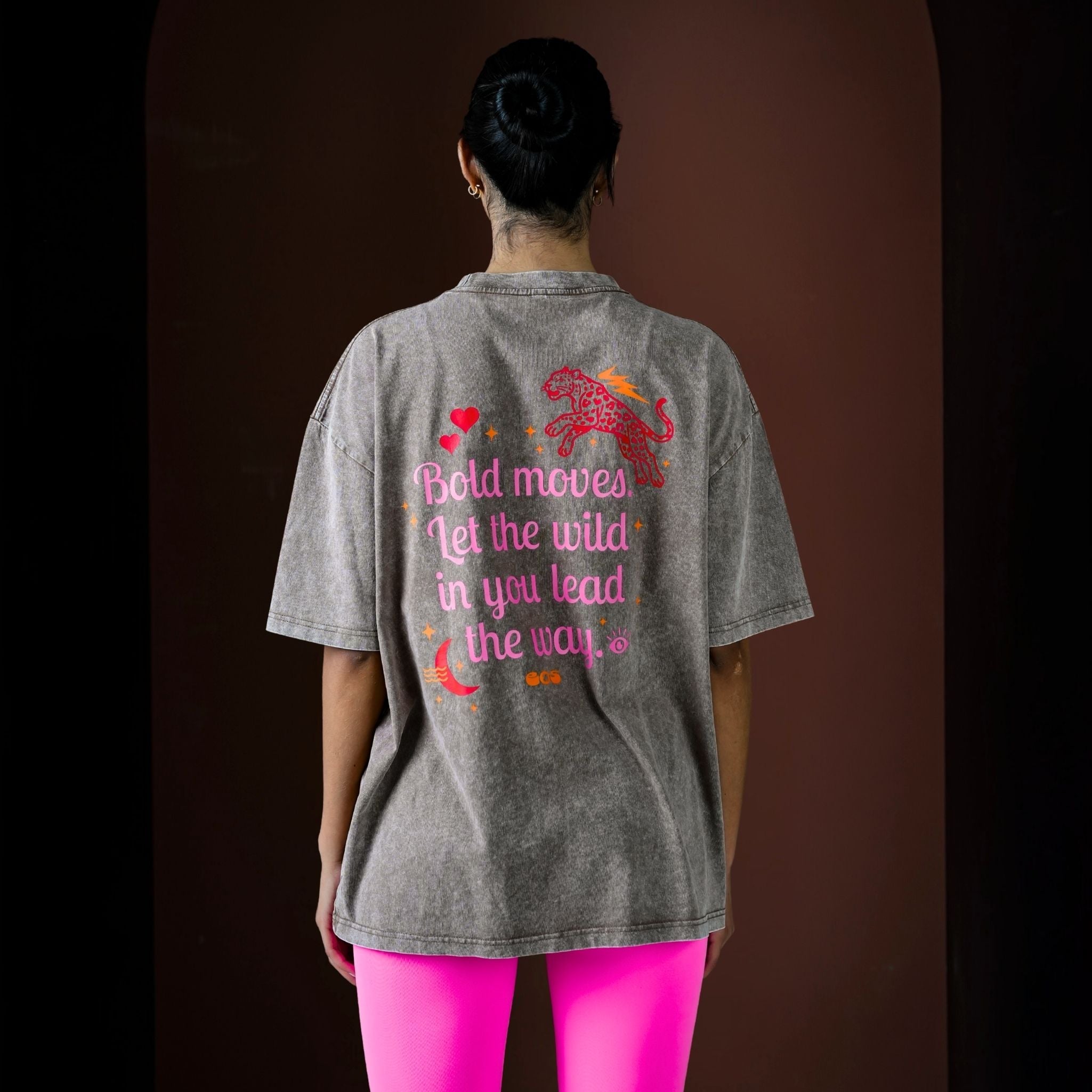 Person wearing a gray t-shirt with colorful text and graphics on the back, standing against a dark background.