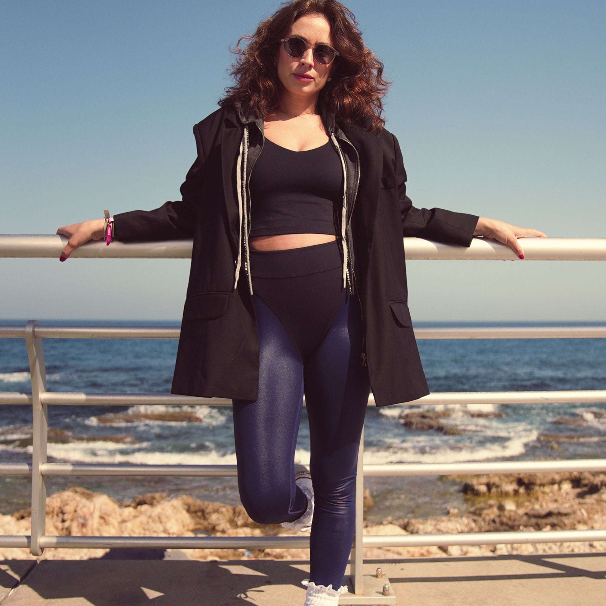 Woman in black coat and leggings standing by a railing with ocean view