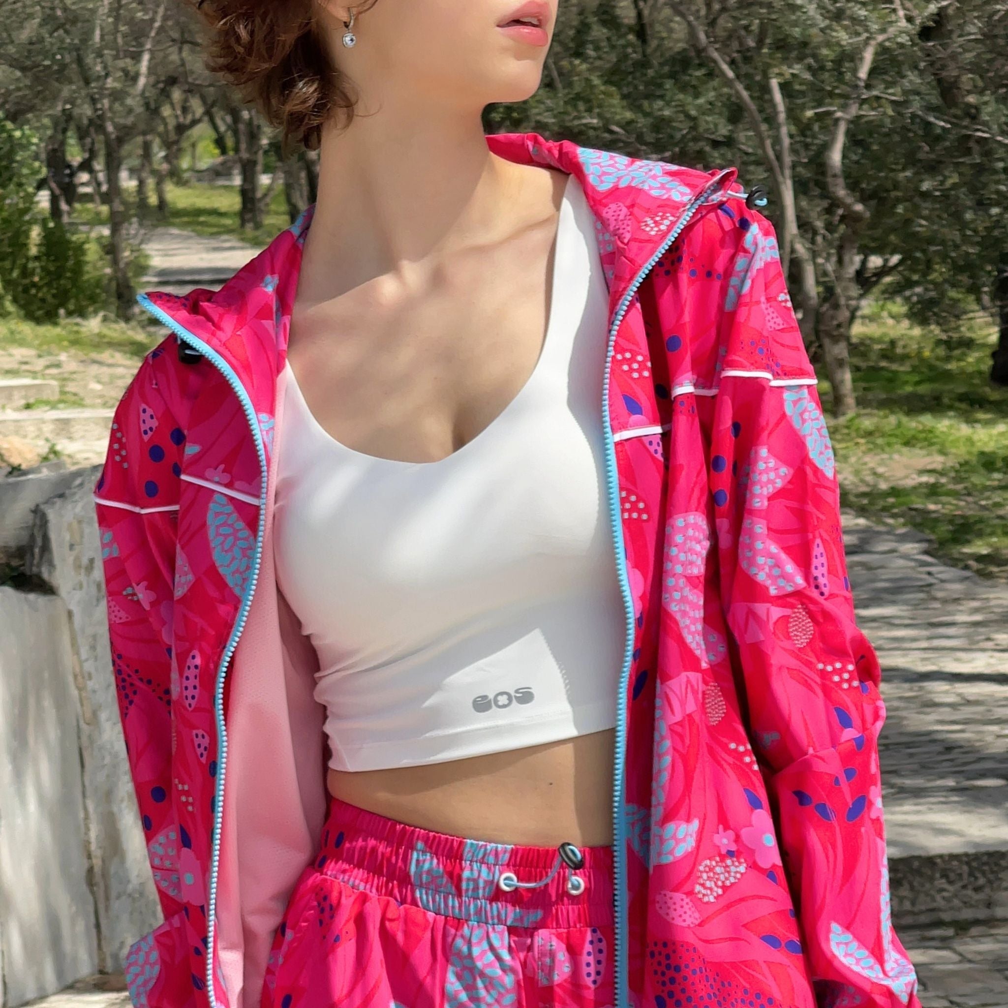 A woman wearing a white bralette with the brand name 'SOS' visible on the front, paired with a bright pink jacket and matching pants with a floral pattern.